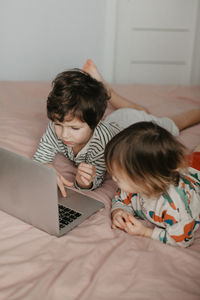 Little brother and sister lie in the bedroom on the bed and look at the laptop. high quality photo