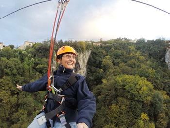 Smiling mature woman hanging on zip line over trees against sky