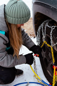 Side view of female wearing knitted hat and gloves and warm sportswear squatting and putting security snow chain on tire wheel of stuck car in snowy countryside