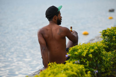 Rear view of shirtless man sitting by sea with beer bottle