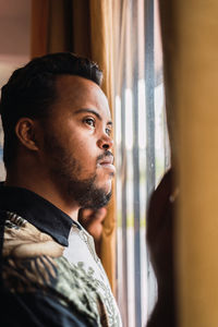 Serious young ethnic man standing near window while looking away pensively in bright house