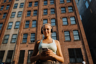 Sportive young blonde woman in fitness clothing wearing headphones and turning music on smartphone