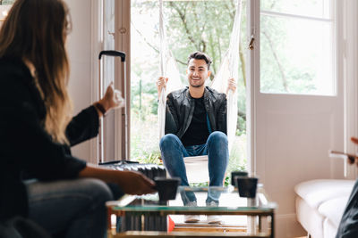 Young woman talking with male friend sitting in swing at living room