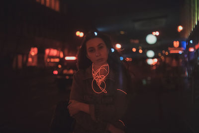 Portrait of young woman with illuminated cable standing on street in city at night