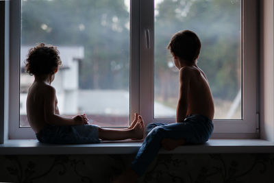 Rear view of real brothers siblings looking through window at home