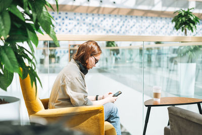 Young teenager girl student in glasses using mobile phone in headphones in yellow chair 