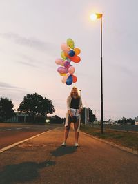 Woman with colorful helium balloons standing on road during sunset