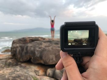 Cropped hand of person photographing woman on rock at beach against sky