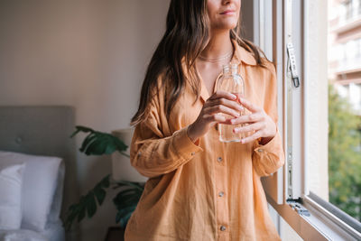 Midsection of young woman holding water bottle by glass window at home