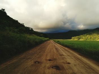 Dirt road leading to grassy field against cloudy sky