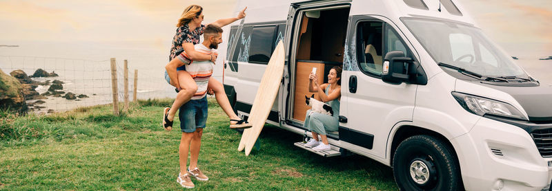 Woman taking photo with mobile of her friends piggybacking next to their camper van during a trip