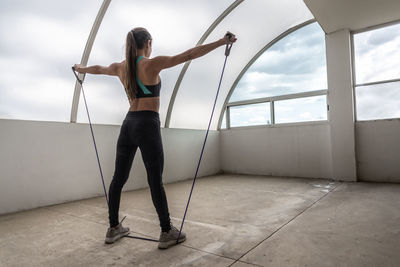 Back view of unrecognizable female athlete in sportswear working out with elastic band while looking forward