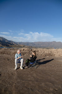 Woman and her grandfather talking while sitting on camping chairs outdoors in nature.