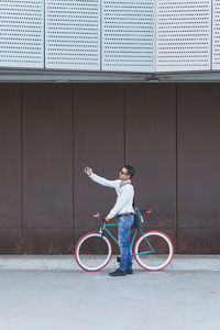 Side view of mid adult man with bicycle taking selfie while standing against building