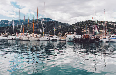 Boats moored at harbor by mountains against cloudy sky