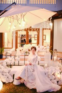 Full length portrait of bride wearing wedding dress sitting against house at night