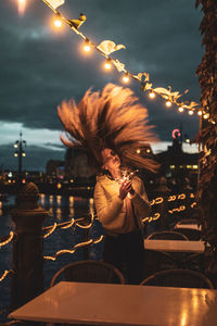 Portrait of smiling young woman tossing hair while holding illuminated lighting equipment at night