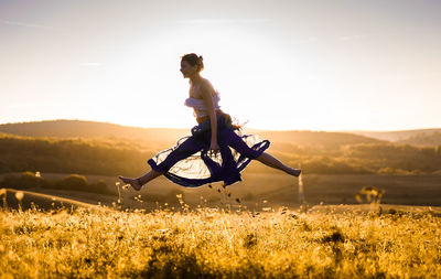 Side view of woman jumping over plants against sky during sunset