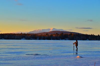 Person drilling at frozen lake against sky during sunset
