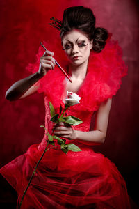 Young woman in red dress holding paintbrush and white rose against black background