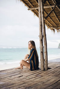 Young woman sitting on shore at beach against sky