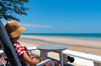 Senior woman sitting on balcony looking at blue sea and white beach in sunny day of summer vacation