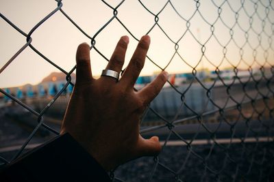 Close-up of hand holding chainlink fence against sunset sky