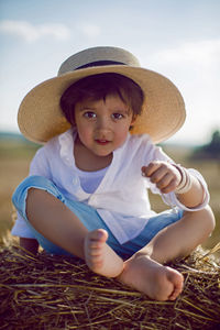 Baby boy in straw hat and blue pants sitting on a haystack in a field in autumn