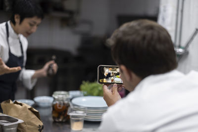 Male colleague filming chef preparing food in kitchen of restaurant