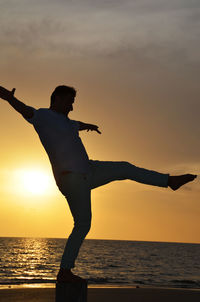 Silhouette man practicing karate at seashore against sky during sunset