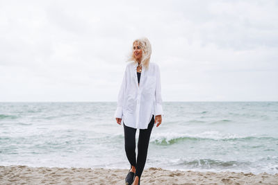 Portrait of elegant blonde woman in white shirt on sand beach at storm sea at windy weater