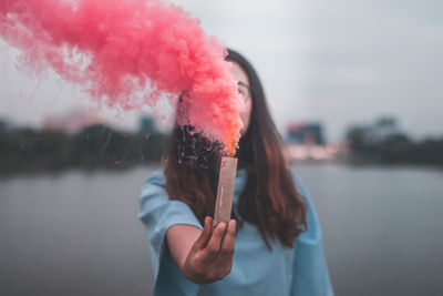 Young woman holding distress flare while standing by lake against sky