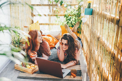 Joyful girls use laptop and drink cocktails spending time together on light decorated terrace 