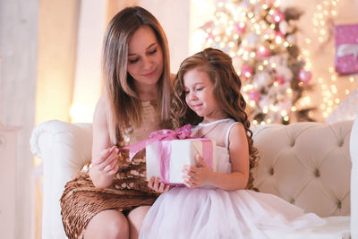 A fasion woman is presenting christmas gift for daughter sitting on the sofa in light living room.