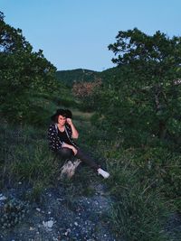 Young woman sitting on field by trees against sky