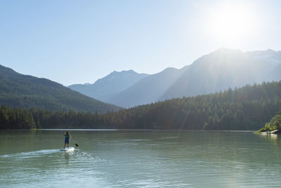 Back view of anonymous female riding sup board on peaceful lake near mountains and forest on sunny day in highlands of british columbia, canada