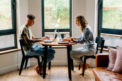 Millennial woman and man work remotely on laptops while sitting at a table in a cozy apartment.