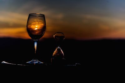 Close-up of wineglass and hourglass against sky during sunset