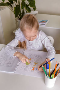 A girl student sits at a desk in the classroom and paints a picture with pencils. 