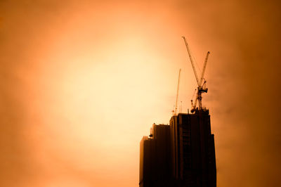 Low angle view of silhouette crane by building against sky during sunset