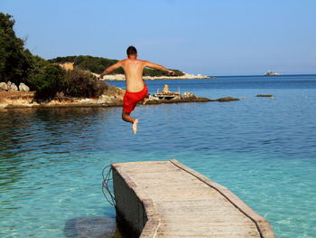 Rear view of shirtless man jumping in sea against sky