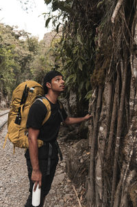 Side view of young man standing by tree in forest