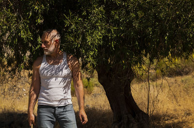 Adult man in white tank top and jeans standing with olive tree in summer