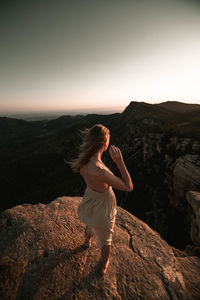 Rear view full length of young woman standing on rock against clear sky during sunset