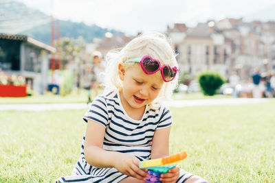 A little blonde girl sitting on the lawn plays with a trendy toy pop it. 