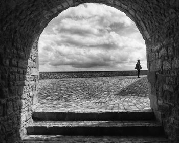 Woman standing on footpath against cloudy sky seen through arch