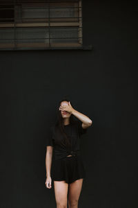 Anonymous trendy female covering eyes while standing on background of black wall of building in city in costa rica