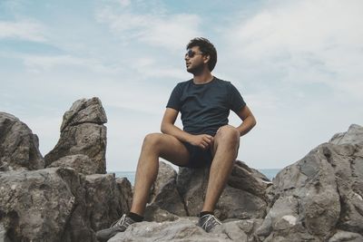 Full length of young man sitting on rocks at beach against sky