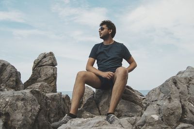 Young man sitting on rock against sky