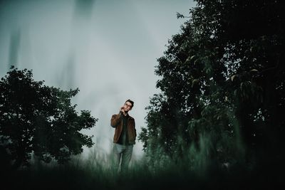 Young man standing by trees against sky
